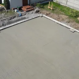 Świeżo wylany, wyrównany fundament pod budynek z widocznymi śladami po listwie wibracyjnej, obrzeżami oraz przygotowanym terenem wokół.