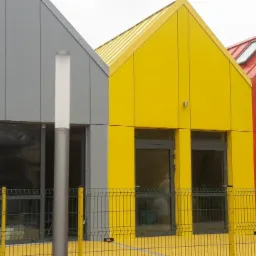 Trzy budynki o nowoczesnej architekturze z elewacjami w kolorach szarym, żółtym i czerwonym, każdy z dwuspadowym dachem w odpowiadającym kolorze, widoczne okna i drzwi, na pierwszym planie metalowe...