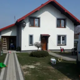 Dom jednorodzinny po malowaniu elewacji na biało z szarymi obramowaniami okien i czerwoną dachówką, widoczne puszki z farbą i drabina.