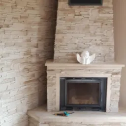 Wykończony kominek obłożony kamieniem dekoracyjnym, z czarnym wkładem kominkowym, ozdobiony figurką anioła i narzędziami na blacie, z widoczną ścianą wykończoną tym samym kamieniem.