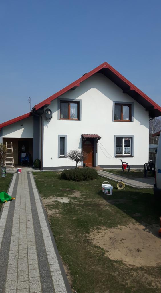 Dom jednorodzinny po malowaniu elewacji na biało z szarymi obramowaniami okien i czerwoną dachówką, widoczne puszki z farbą i drabina.