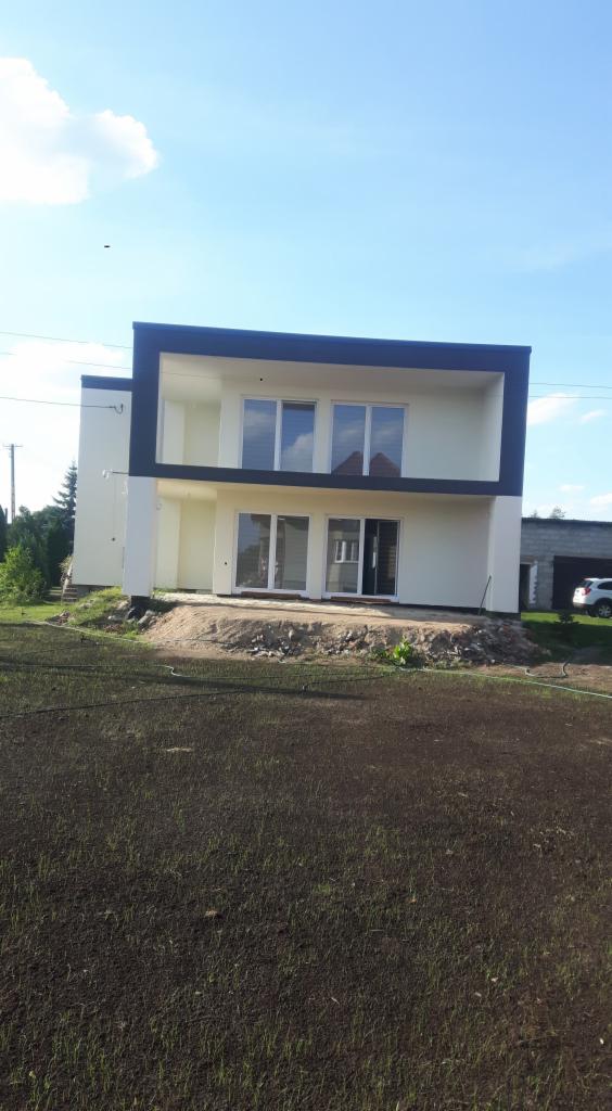 Nowoczesny dwupiętrowy dom z płaskim dachem i jasną elewacją, czarne obramowanie okien i balkonu, teren wokół domu w trakcie zagospodarowywania, widoczny garaż i samochód w tle.