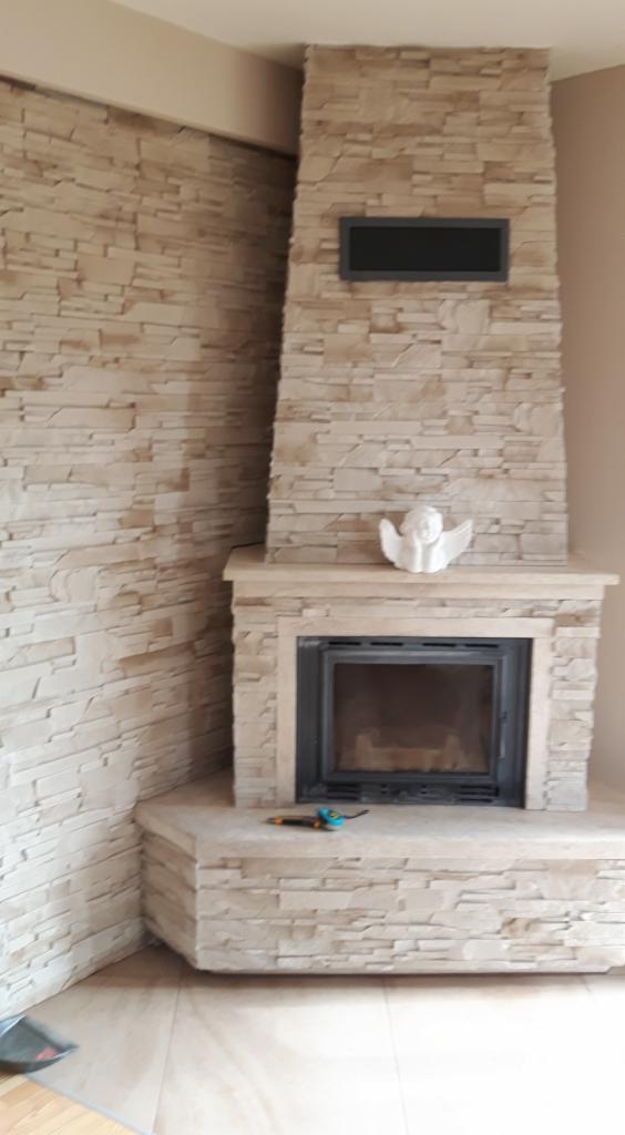 Wykończony kominek obłożony kamieniem dekoracyjnym, z czarnym wkładem kominkowym, ozdobiony figurką anioła i narzędziami na blacie, z widoczną ścianą wykończoną tym samym kamieniem.