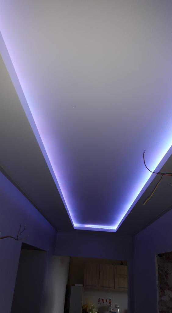 Nowoczesny sufit podwieszany z oświetleniem LED w kolorze fioletowym, widoczne fragmenty instalacji elektrycznej i mebli kuchennych w tle.