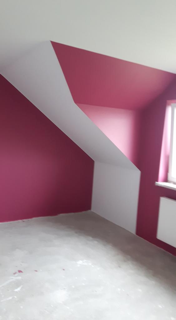 Pomalowane na biało i malinowo wnętrze poddasza ze skosami, widok na okno z grzejnikiem i surową posadzkę.