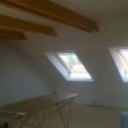 Poddasze po szpachlowaniu i malowaniu na biało, widoczne dwa okna dachowe, surowe belki stropowe i metalowy stół warsztatowy.