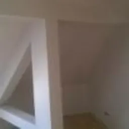 Wnętrze w trakcie wykańczania: białe ściany, widoczne łączenia płyt gipsowo-kartonowych przed szpachlowaniem, surowe oświetlenie podkreśla nierówności powierzchni.