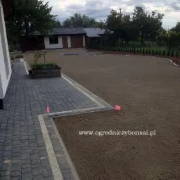 Świeżo ułożona szara kostka brukowa z jasną obwódką granicząca z przygotowanym terenem pod trawnik, w tle budynek gospodarczy i rząd małych drzewek.