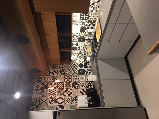 Wnętrze kuchni z wzorzystymi, czarno-białymi płytkami na ścianie, drewnianą zabudową sufitową z oświetleniem punktowym i szarymi szafkami kuchennymi.