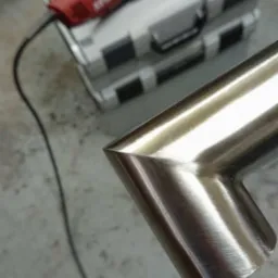 Zespawany pod kątem fragment rury ze stali nierdzewnej, widoczny szlif na powierzchni, w tle narzędzie do obróbki metalu i skrzynka narzędziowa.