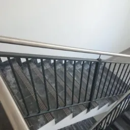 Schody z czarnymi metalowymi balustradami i srebrną, zaokrągloną poręczą, widok z góry na klatce schodowej wykończonej szarą płytką.