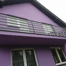 Balkon z prostą, poziomą, czarną balustradą na fioletowym budynku z widocznymi oknami.