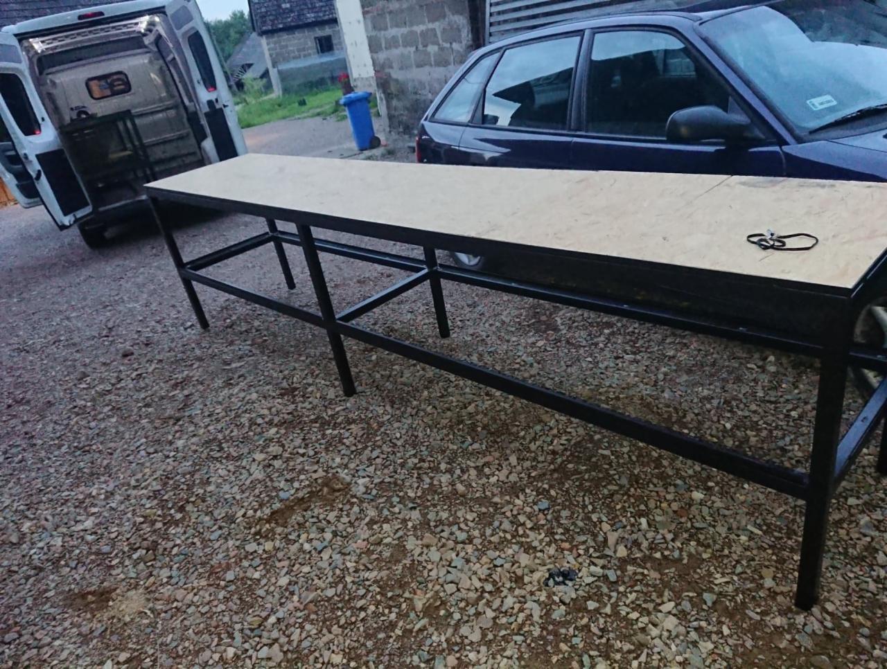 Długi, metalowy stół z drewnianym blatem, ustawiony na żwirowym podłożu, z widocznym samochodem osobowym i dostawczym w tle.
