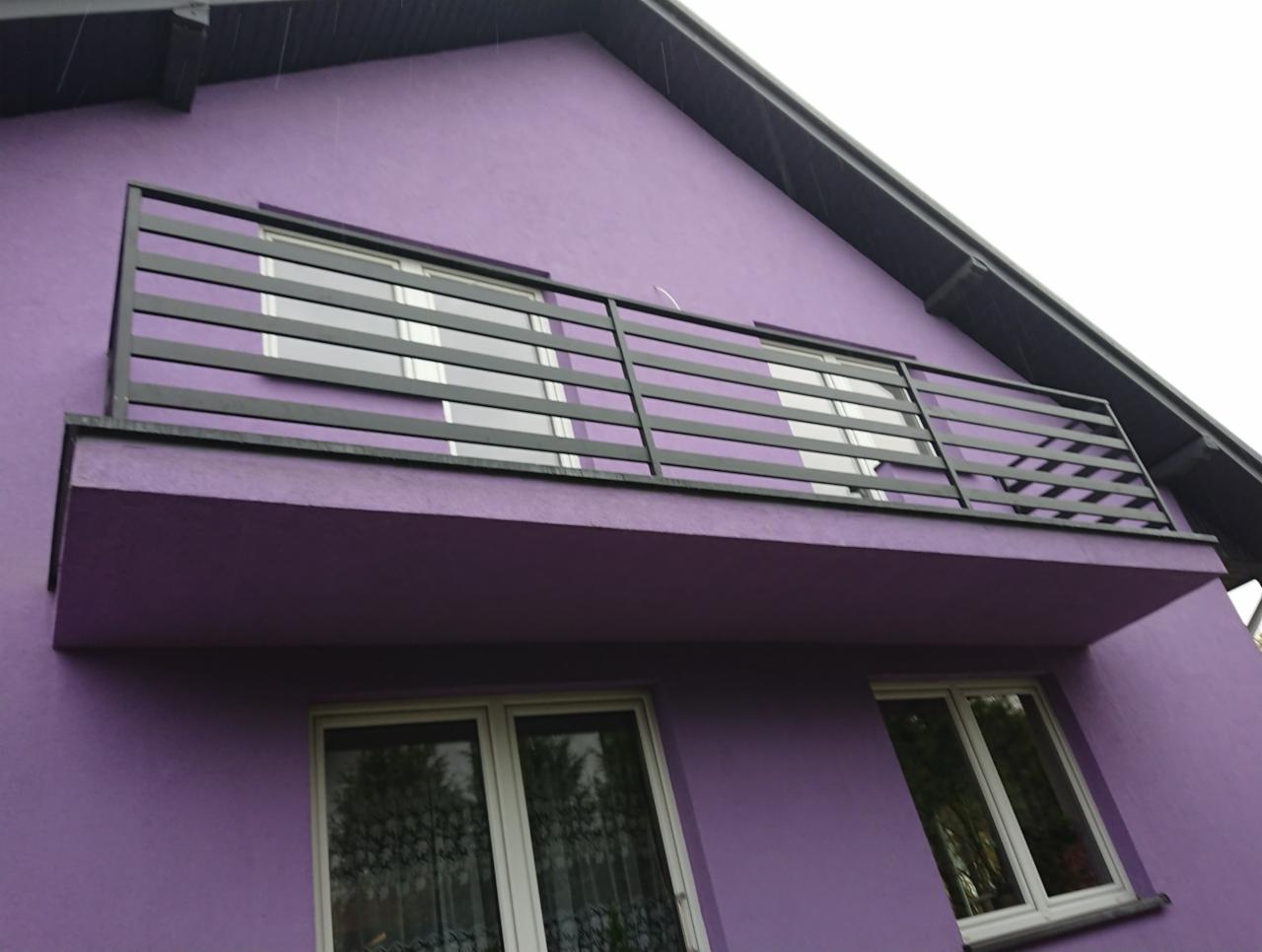 Balkon z prostą, poziomą, czarną balustradą na fioletowym budynku z widocznymi oknami.