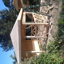 Konstrukcja drewnianej altany w trakcie budowy, widoczne elementy kratowe, drabina oparta o budowlę, w tle drzewo iglaste i fragment innej konstrukcji.