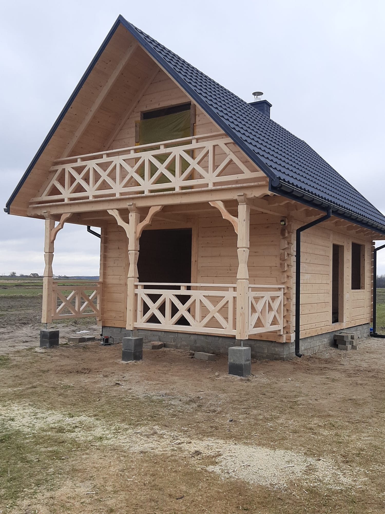 Surowy, dwukondygnacyjny dom szkieletowy z jasnego drewna, z balkonem i czarnym dachem, osadzony na betonowych fundamentach. Widok z zewnątrz na tle pola.