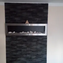 Ściana z ciemnej okładziny imitującej kamień, z wnęką w stalowej ramie wypełnioną białymi kamieniami dekoracyjnymi i drewnianą ozdobą w kształcie piramidy na górnej krawędzi ramy.