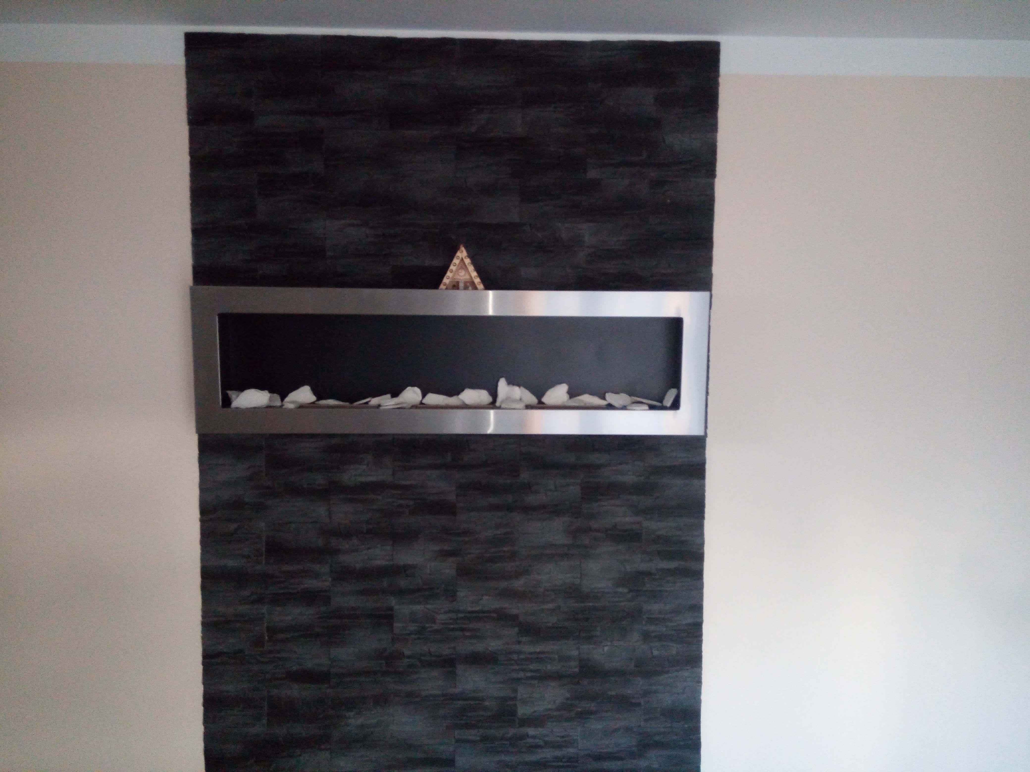 Ściana z ciemnej okładziny imitującej kamień, z wnęką w stalowej ramie wypełnioną białymi kamieniami dekoracyjnymi i drewnianą ozdobą w kształcie piramidy na górnej krawędzi ramy.