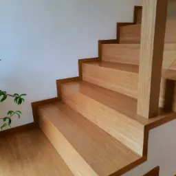 Jasne drewniane schody z minimalistyczną balustradą i rośliną w tle, widok z dołu ukazujący strukturę drewna.