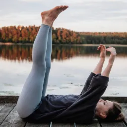 Kobieta w szarym dresie wykonuje ćwiczenie z uniesionymi nogami na drewnianym pomoście nad jeziorem o zachodzie słońca, jesienne drzewa w tle.