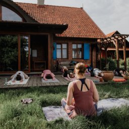 Jogadom - Grupa osób ćwiczy na matach w plenerze przed rustykalnym budynkiem z drewnianymi elementami, trawa w pierwszym planie, widok zza pleców siedzącej kobiety.
