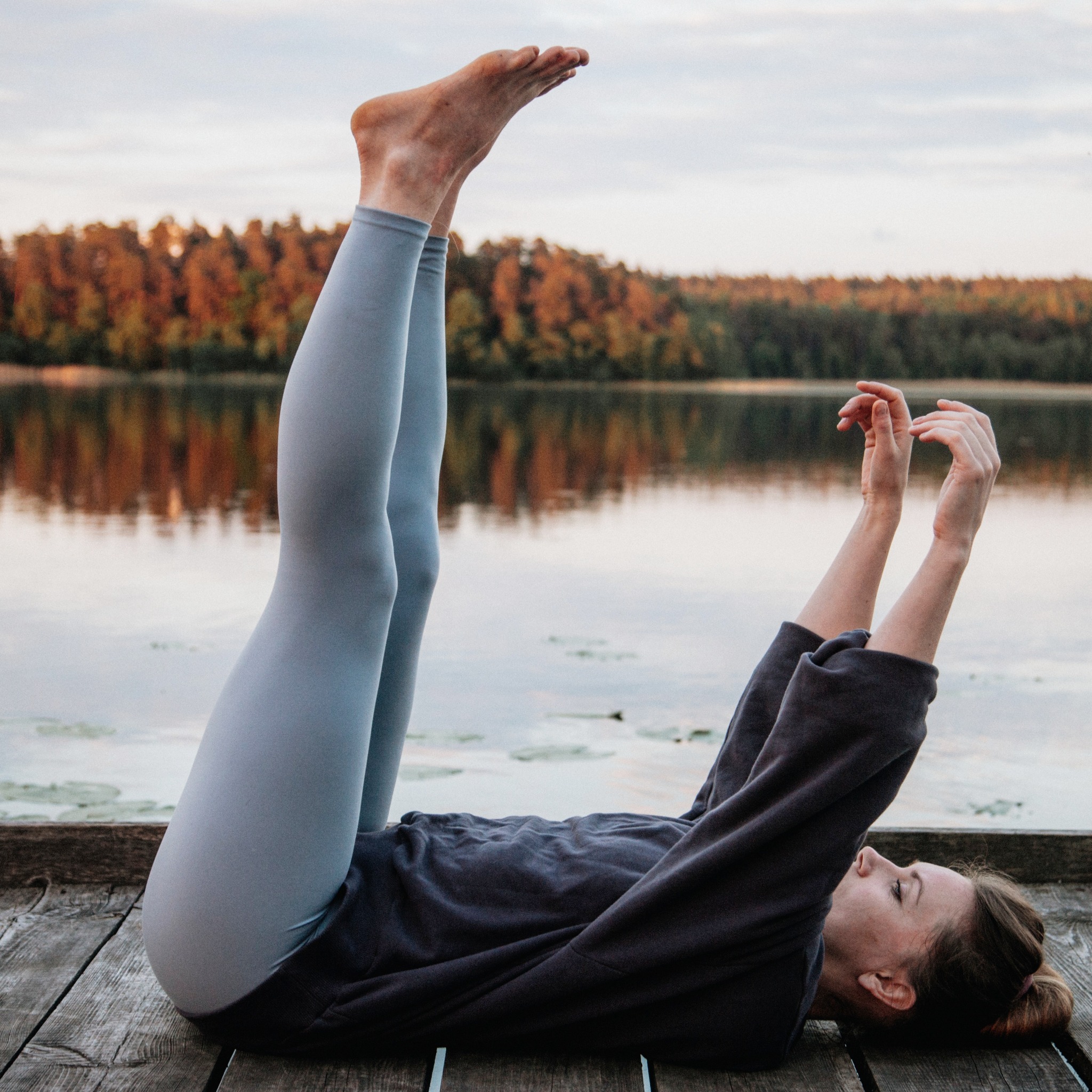 Kobieta w szarym dresie wykonuje ćwiczenie z uniesionymi nogami na drewnianym pomoście nad jeziorem o zachodzie słońca, jesienne drzewa w tle.