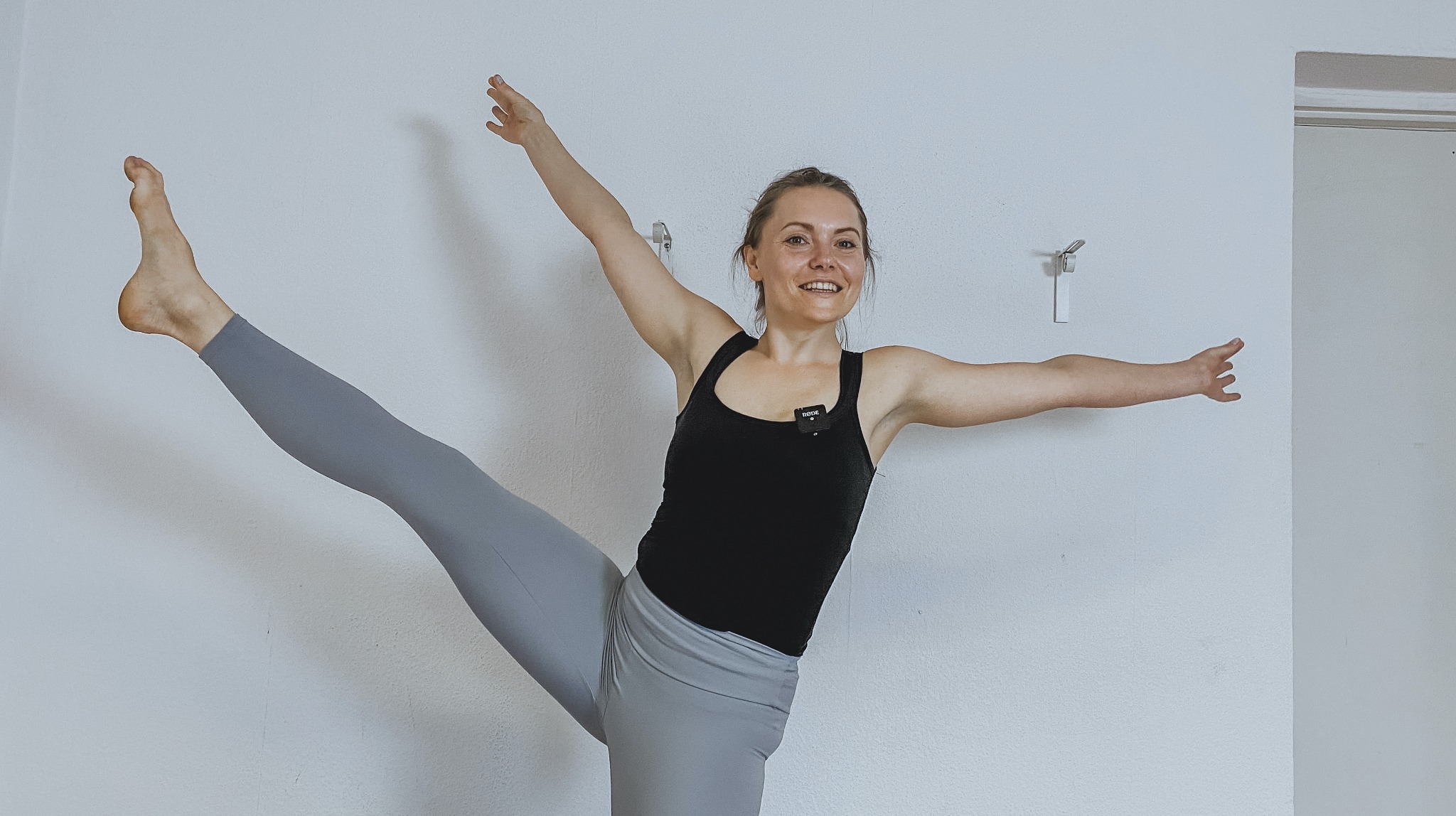 Uśmiechnięta kobieta w czarnym topie i szarych legginsach, wykonująca pozycję stojącą z uniesioną nogą, mikrofon przypięty do ubrania, białe tło