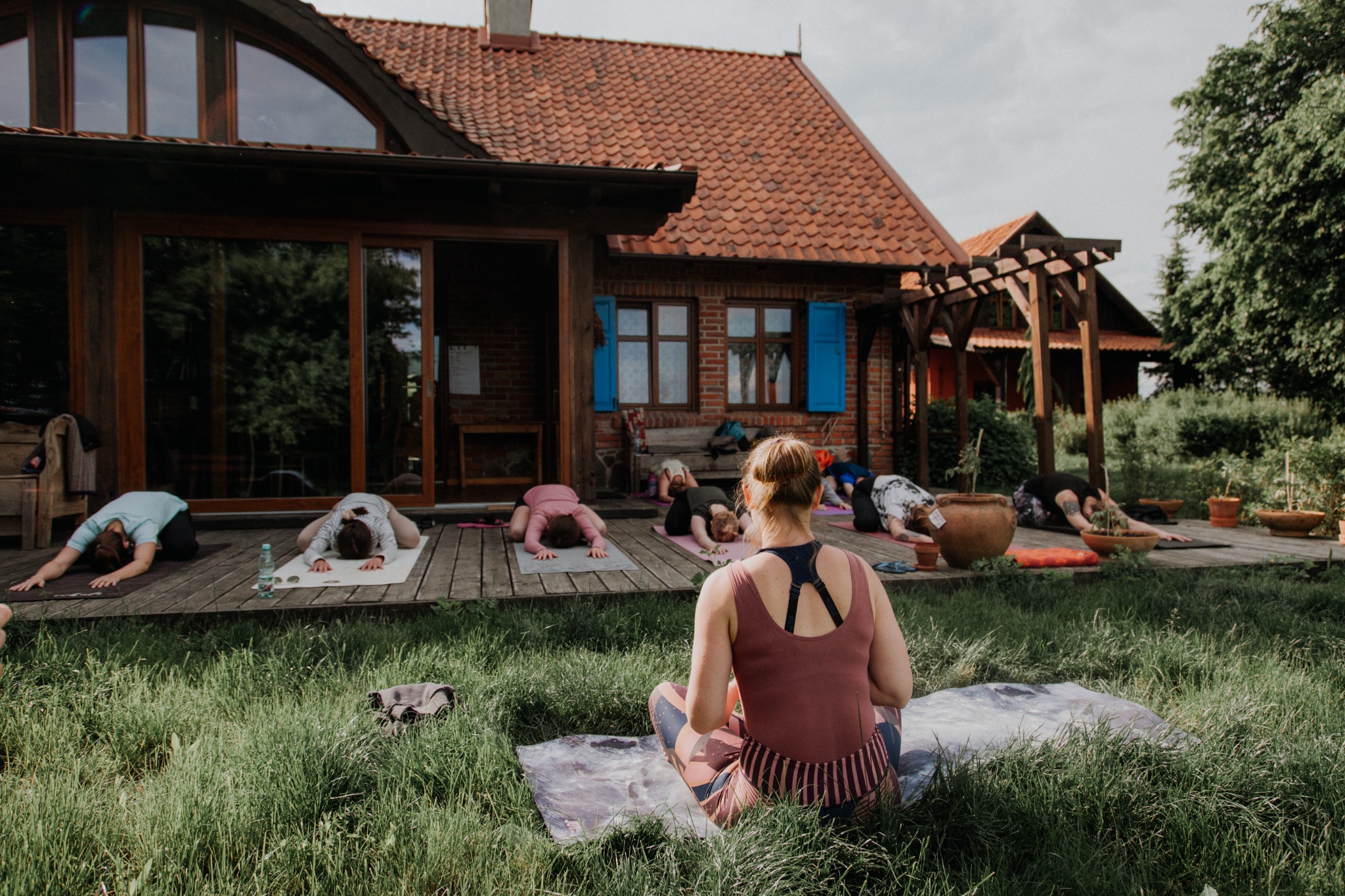 Grupa osób ćwiczy na matach w plenerze przed rustykalnym budynkiem z drewnianymi elementami, trawa w pierwszym planie, widok zza pleców siedzącej kobiety.