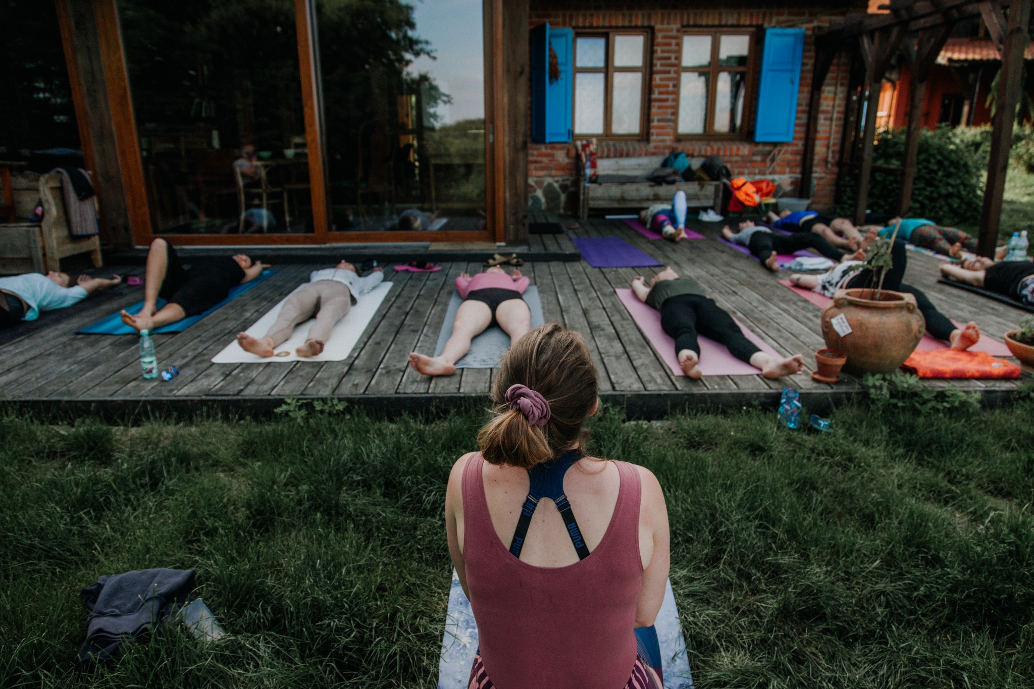Grupa osób relaksuje się na matach na drewnianym tarasie podczas zajęć na świeżym powietrzu, widok z perspektywy osoby siedzącej na macie na trawie, butelki wody obok uczestników.