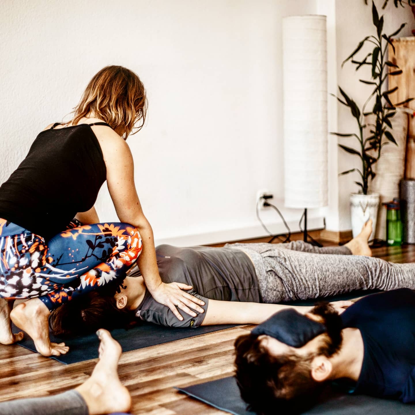 Instruktorka jogi w kolorowych legginsach wykonuje masaż głowy leżącej osobie na macie w jasnym wnętrzu studia, widoczne stopy innych uczestników.