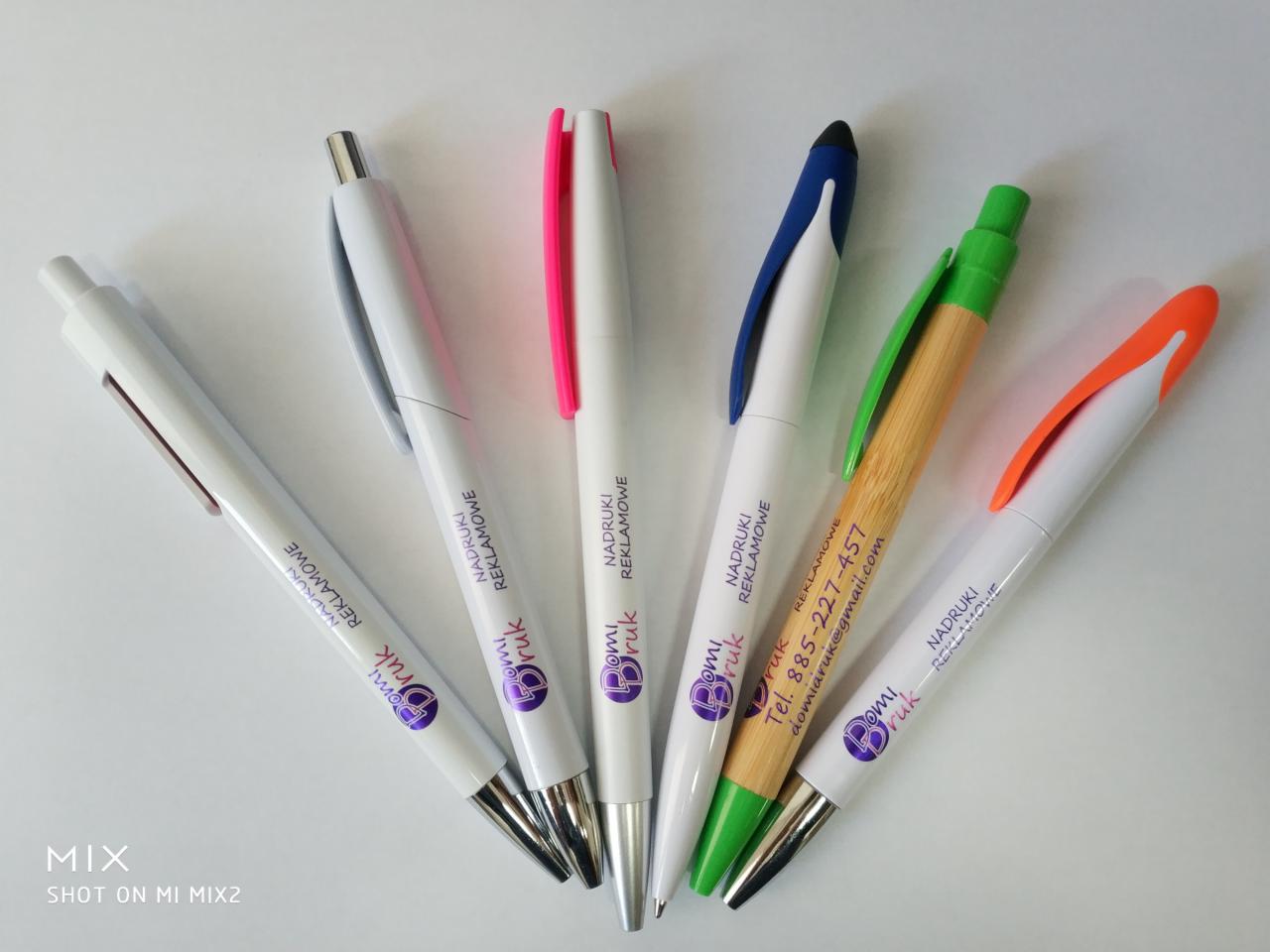 Sześć długopisów reklamowych z nadrukiem firmowym, leżących wachlarzem na białym tle, z różnymi kolorami skuwek: srebrnym, różowym, niebieskim, zielonym (na bambusowym korpusie) i pomarańczowym.