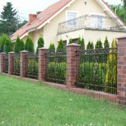 Ozdobne, kute przęsła ogrodzeniowe w kolorze ciemnego brązu, osadzone na niskim cokole z czerwonej cegły i słupkach z tego samego materiału, na tle zadbanego trawnika i domu z balkonem.