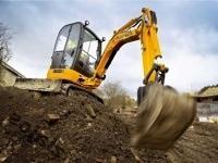 Żółta koparka JCB na placu budowy, łyżka zagłębiona w ziemi, w tle budynki mieszkalne i pochmurne niebo.