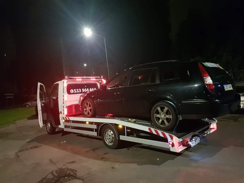 Czarny samochód kombi załadowany na białą lawetę z otwartymi drzwiami kierowcy, widok nocny pod latarnią.