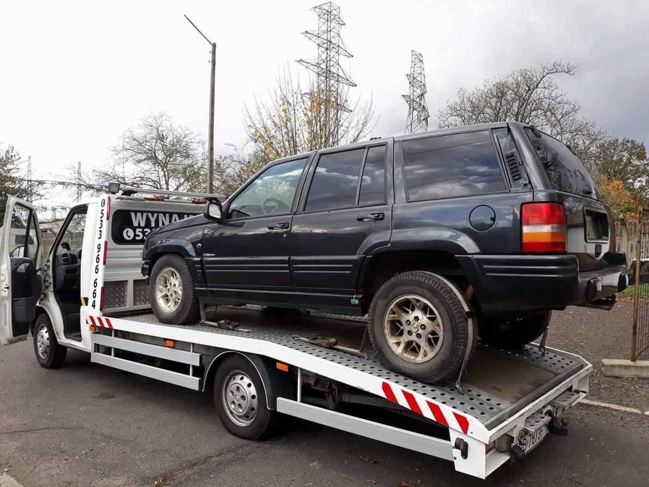 Czarny Jeep Grand Cherokee transportowany na białej lawecie z otwartymi drzwiami kabiny, widoczne słupy wysokiego napięcia w tle.