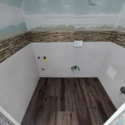 Wnętrze łazienki w trakcie prac wykończeniowych: brązowe panele podłogowe imitujące drewno, jasne płytki ścienne z dekoracyjnym pasem mozaiki, surowy sufit z widocznymi śladami prac, instalacja...