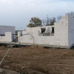 Wznoszenie ścian parterowego domu z białych bloczków na fundamencie, widoczne zbrojenia okienne, rury instalacyjne i teren budowy z ziemią.