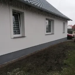 Boczny widok ocieplonego domu z białym tynkiem i szarą podmurówką, z dekoracyjnym oświetleniem zwisającym z dachu, widoczne okna z roletami dzień-noc.