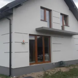 Dom jednorodzinny z jasną elewacją, szarymi akcentami i brązowymi ramami okiennymi, widok z boku na ogród z fragmentem szklarni.