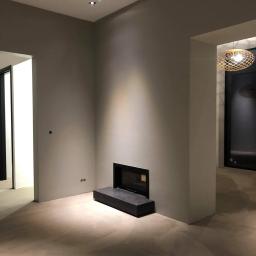 SenS- Wykończenia - Wnętrze po pracach wykończeniowych: gładkie, szare ściany, wbudowany kominek z czarną obudową, trzy punktowe lampy sufitowe i ozdobna lampa wisząca w tle.