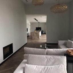 SenS- Wykończenia - Jasnoszary salon z parkietem w jodełkę, minimalistycznym kominkiem, otwartą kuchnią w tle i ozdobnymi lampami sufitowymi.