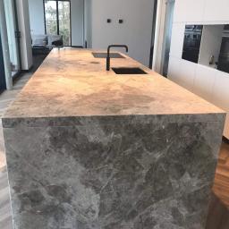 SenS- Wykończenia - Wyspa kuchenna z blatem z szarego marmuru z czarną armaturą, widok na salon i okno w tle, parkiet w jodełkę na podłodze.