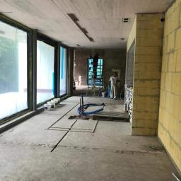 SenS- Wykończenia - Wnętrze w stanie deweloperskim z surowym betonowym stropem, widoczne puszki z farbą przy oknie, instalacje wyprowadzone z podłogi, pracownik na drabinie w oddali oraz ściana z bloczków.