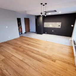 Wnętrze mieszkania po remoncie z nową drewnianą podłogą, szarą ścianą i czarną ścianą z oświetleniem punktowym, widoczne płytki dekoracyjne i okno z folią ochronną.