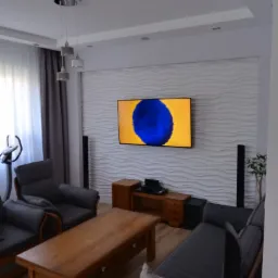 Nowoczesny salon z teksturowaną ścianą 3D, telewizorem, szarą sofą i drewnianym stolikiem kawowym oraz lampami wiszącymi.