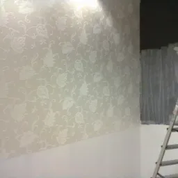 Ściana pokryta jasną tapetą z wytłaczanym, roślinnym wzorem, widoczna drabina aluminiowa i fragment ciemnej ściany w rogu pomieszczenia.