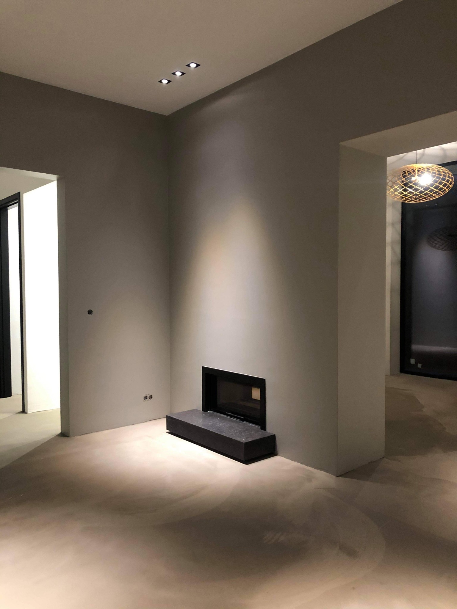 Wnętrze po pracach wykończeniowych: gładkie, szare ściany, wbudowany kominek z czarną obudową, trzy punktowe lampy sufitowe i ozdobna lampa wisząca w tle.