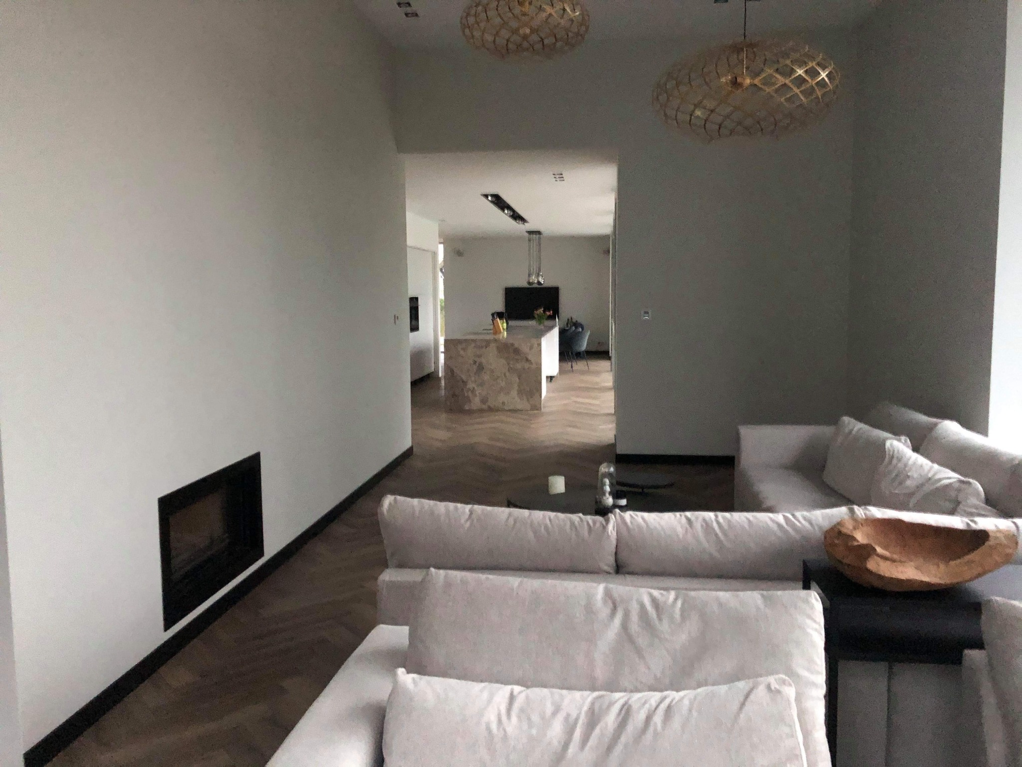 Jasnoszary salon z parkietem w jodełkę, minimalistycznym kominkiem, otwartą kuchnią w tle i ozdobnymi lampami sufitowymi.