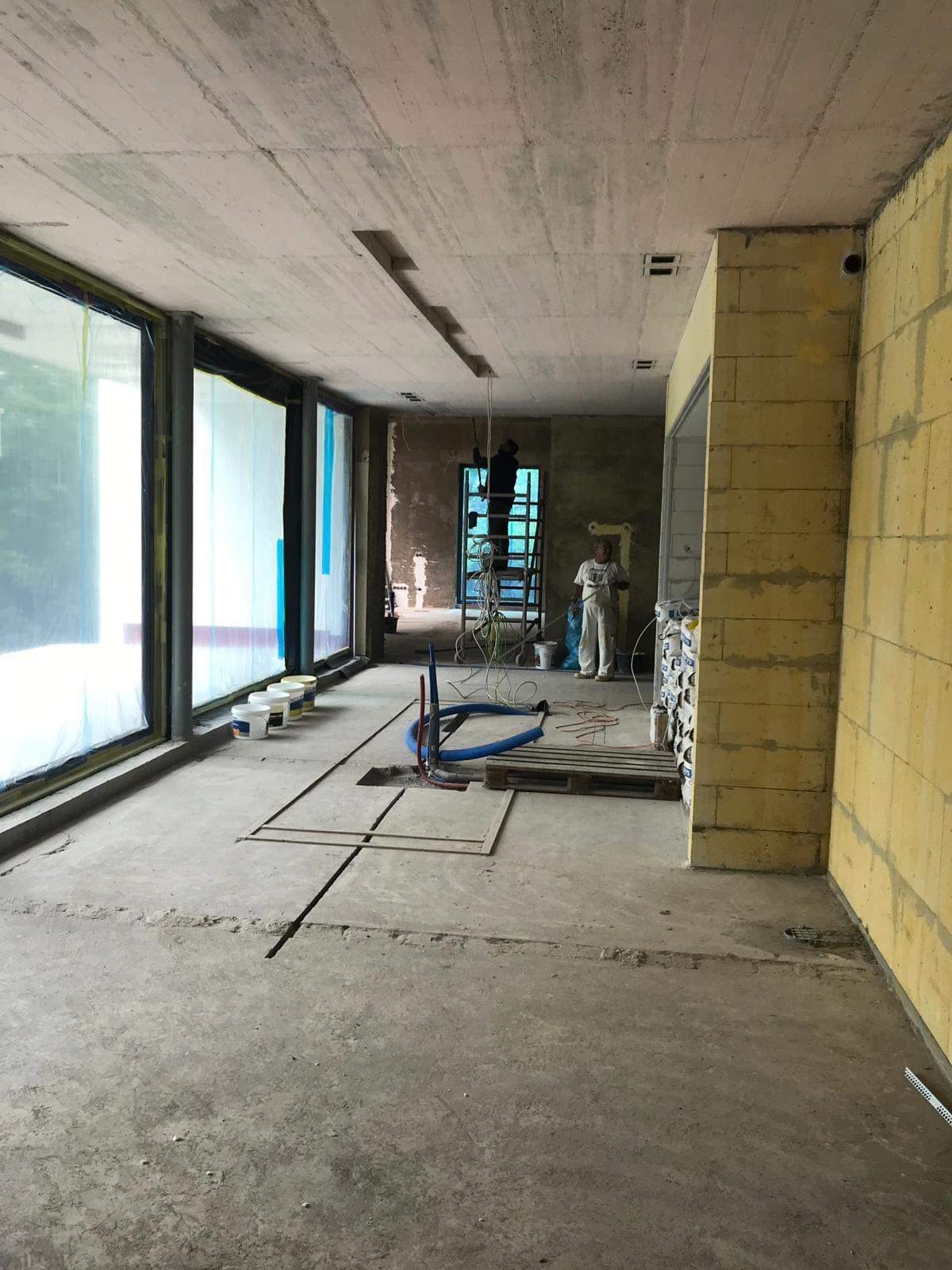 Wnętrze w stanie deweloperskim z surowym betonowym stropem, widoczne puszki z farbą przy oknie, instalacje wyprowadzone z podłogi, pracownik na drabinie w oddali oraz ściana z bloczków.