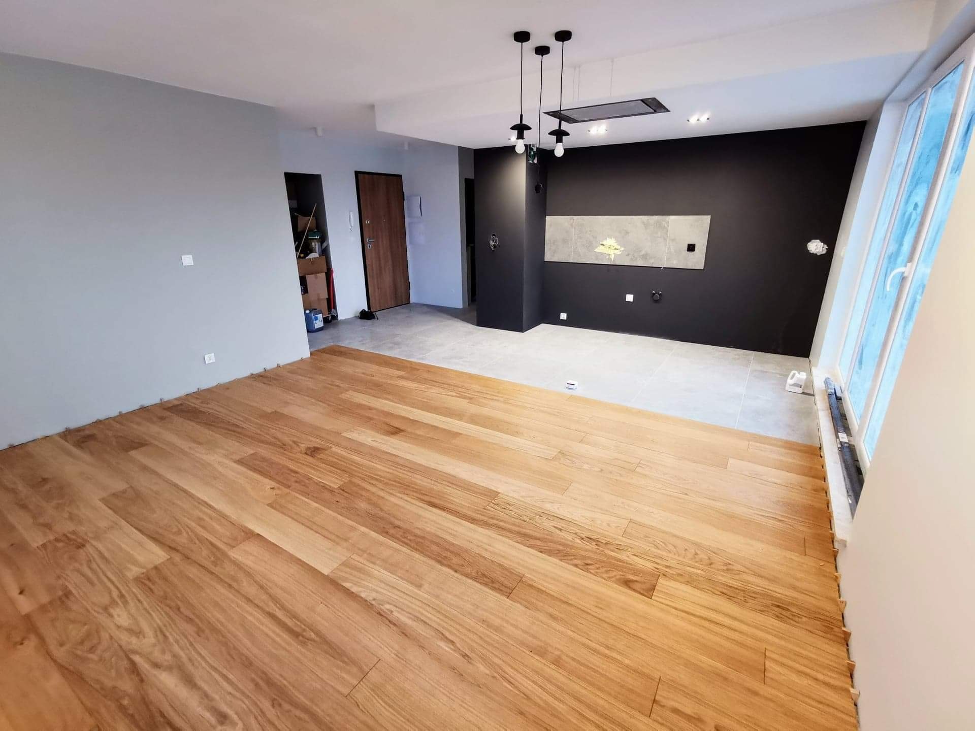 Wnętrze mieszkania po remoncie z nową drewnianą podłogą, szarą ścianą i czarną ścianą z oświetleniem punktowym, widoczne płytki dekoracyjne i okno z folią ochronną.